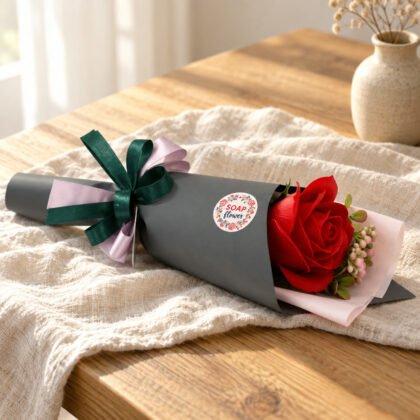 Ramo de uma rosa vermelha de sabão em papel cinzento com autocolante SOAP flower e laços verde e lilás, sobre linho numa mesa de carvalho