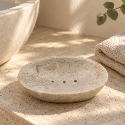 Saboneteira oval em mármore creme numa bancada de travertino ao lado do lavatório
