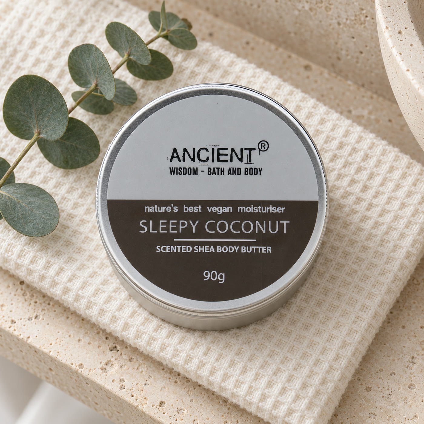 Lata de manteiga corporal Sleepy Coconut da Ancient Wisdom, 90 g, sobre uma toalha de favo creme numa prateleira de travertino