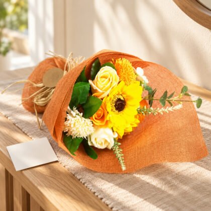 Ramo de flores de sabão laranja e amarelo com girassol, embrulhado em papel laranja com ráfia, numa mesa de entrada com cartão em branco