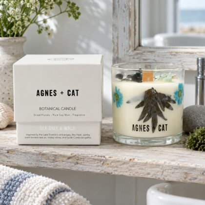 Vela botânica Agnes + Cat Sal Marinho e Musgo com flores secas e a caixa, numa prateleira de madeira de uma casa de banho junto ao mar, com uma pedra e musgo