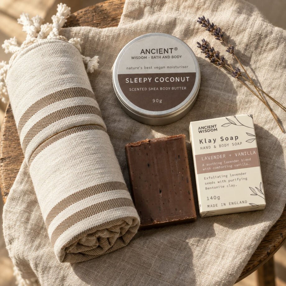 Conjunto Um cuidado só teu visto de cima: toalha de spa às riscas, manteiga corporal Sleepy Coconut e sabonete Klay Soap lavanda e baunilha da Ancient Wisdom sobre linho num banco de madeira